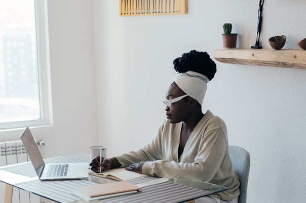 A Black woman sitting at a desk with a laptop focused on her work. She is wearing glasses and a white shirt. She is wearing a headband with her hair pulled up in a bun. The desk is against a wall with a window and above the desk is a shelf with a potted plant. There is also a decorative orange painting on the wall
