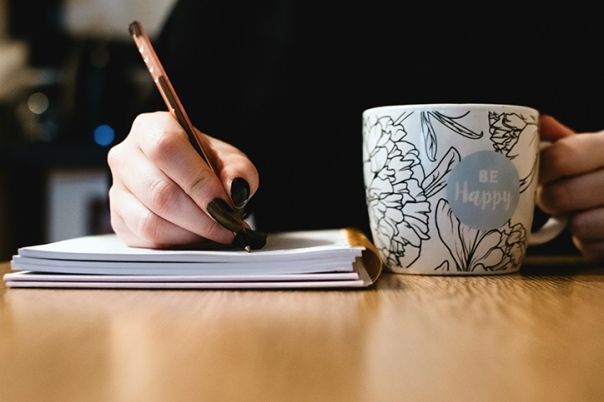 A person’s hand holding a pen and writing in a notebook. Next to the notebook is a mug with floral designs with the words saying be happy written on it.